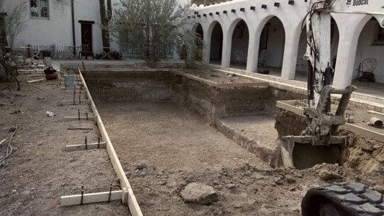An emptied pool void beside a white arched colonnade, string lines set and the excavator arm reaching in.