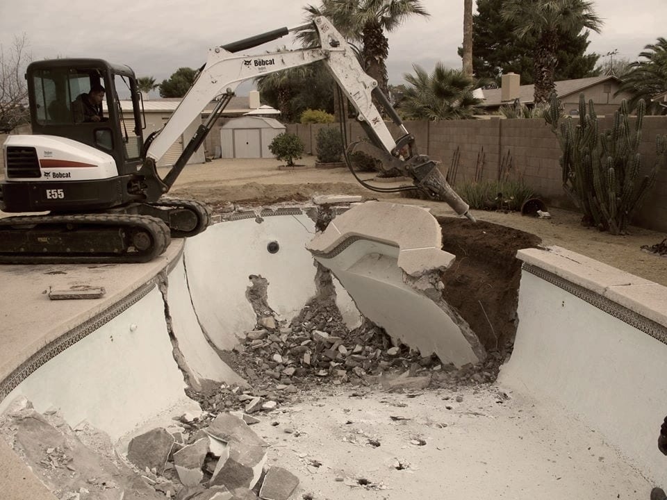 An excavator with its bucket down inside a pool, the shell cracked open and rubble in the bottom.